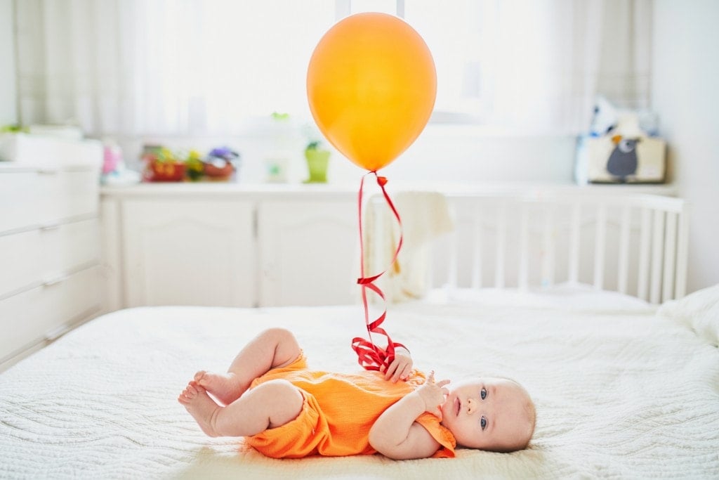 Adorable baby girl in orange romper suit lying on bed in nursery with colorful balloon.
