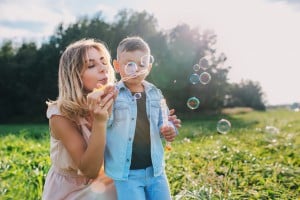 A woman and a child are outdoors in a grassy field. The woman is blowing bubbles while the child watches, holding the bubble bottle. Trees and a clear sky are visible in the background. It's one of those little things parents do that turn moments into cherished memories.