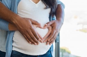 Two people standing close together with their hands forming a heart shape over the pregnant belly of the person in front, capturing a tender moment perfect for Mother's Day while you're expecting. The person behind is wearing a light blue shirt, and the person in front wears a white shirt and a light blue cardigan.