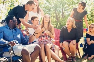 A group of people are gathered outdoors, sitting and standing around red Adirondack chairs. They are laughing and interacting with each other like dads at a baby shower. The setting appears to be a sunny day with green trees in the background.