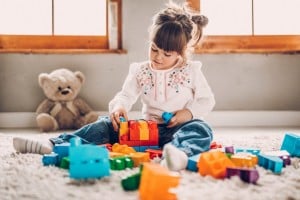 Young child sits on a carpet assembling colorful plastic building blocks, with a toy teddy bear in the background near a window.