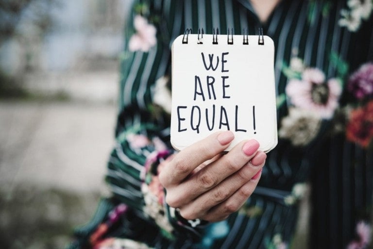 A person in a floral-striped blouse holds a small spiral-bound notebook with the handwritten message "WE ARE EQUAL!" The background appears to be outdoors, and the person's fingernails are painted pink. This powerful image beautifully captures the spirit of Women's Equality Day.