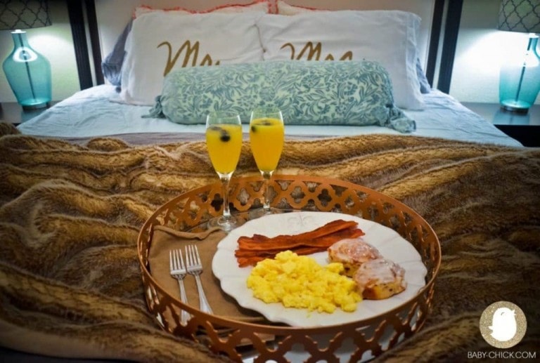 A tray with breakfast items, including scrambled eggs, bacon, and pastries, is placed on a bed with brown fur-like blankets. The tray also holds two mimosas in champagne flutes. In the background, there is a bed with 