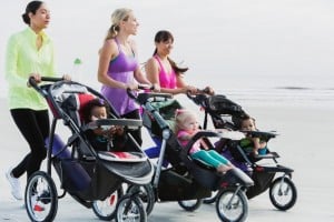 Four mom friends are jogging on a beach while pushing strollers with babies. Dressed in athletic clothing, they chat and laugh under an overcast sky. The strollers, equipped with large wheels suitable for jogging, make it easy to enjoy their workout together.