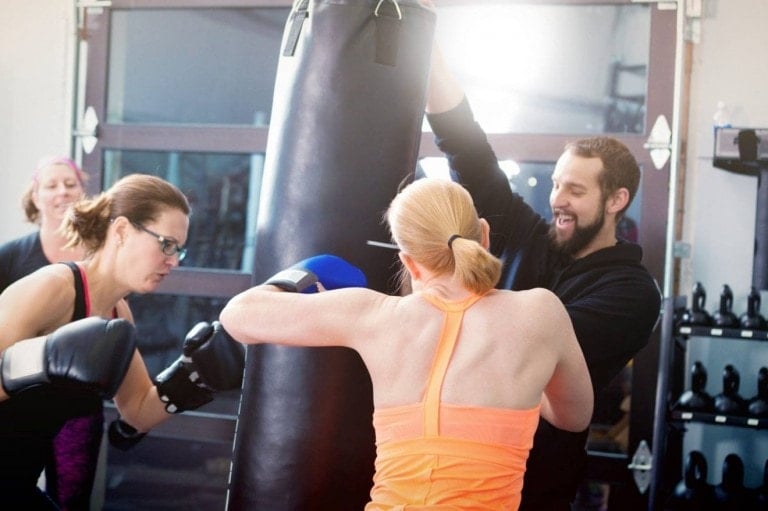 Three individuals exercise in a gym. A man, likely a trainer, holds a punching bag while two women, wearing boxing gloves, engage in boxing exercises—perfect for picking up self defense tips for women. The background includes gym equipment and another person watching. The setting is bright, with light entering from the left.
