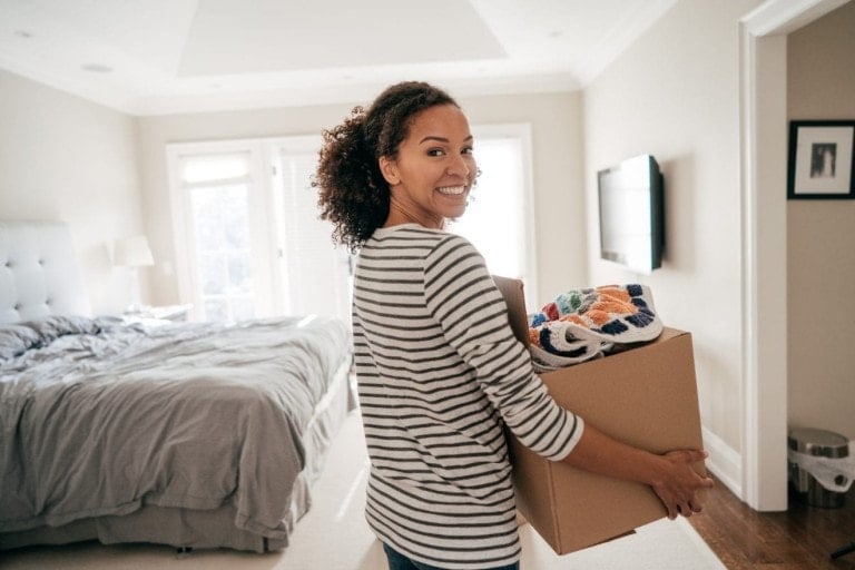 Woman holding a box full of things as she walks through her room.