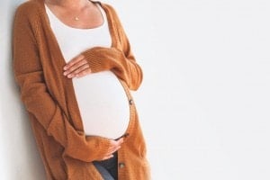 A pregnant woman stands against a white wall, cradling her belly with both hands. She is wearing a white tank top under an open, long brown cardigan. The image is cropped to show her from her neck to her thighs, capturing the tender moment while beautifully illustrating what not to say to a pregnant person.