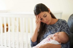 A woman with long dark hair and a blue patterned top holds a sleeping baby in her lap, sitting near a white crib. She rests her head in her hand, appearing tired or stressed, possibly experiencing the baby blues.