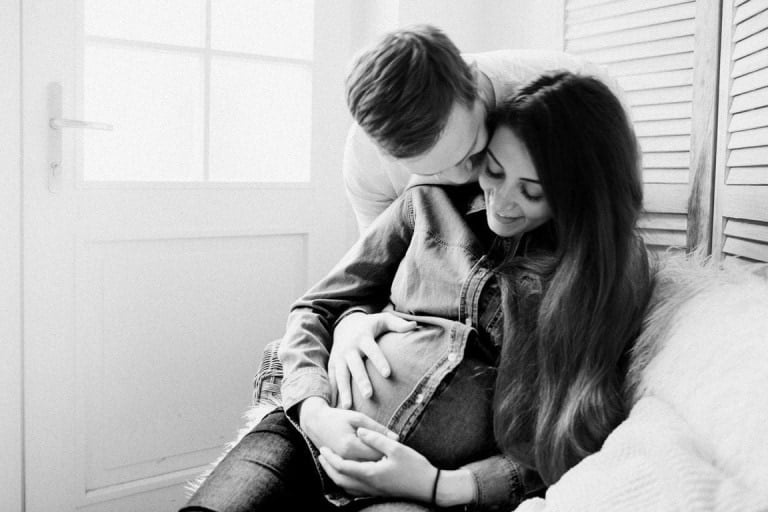 A man and a pregnant woman sit together on a sofa. The man is behind the woman, leaning forward to kiss her on the temple, while both of his hands rest on her pregnant belly, subtly defining their parenting roles. The woman smiles and holds her belly with both hands. The image is in black and white.