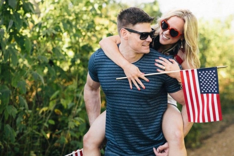 A man wearing sunglasses and a striped T-shirt gives a piggyback ride to a woman also in sunglasses, holding an American flag. They are outdoors with green foliage in the background, letting freedom ring with each smile. She is beaming, and they both appear to be enjoying themselves.