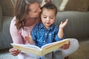 A woman wearing a pink top is sitting closely behind her only child, who is dressed in a blue shirt and gray pants, as they read a book together on the floor. The child, gesturing with one hand while looking at the book, brings a smile to the woman's face.