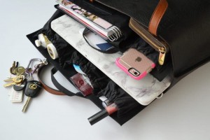 An open black bag with brown handles is displayed, revealing its contents. Inside the entrepreneur's purse are a set of keys, a pink iPhone with a black case, a laptop, a notebook, lipstick, and other personal items. The women entrepreneur purse has multiple compartments for organization.