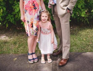 A young girl, often called a threenager, stands in between two adults, holding their hands. She is wearing a white dress with a floral pattern and sandals. The adult on the left is in a colorful dress and platform sandals, while the adult on the right is in a beige suit and brown shoes.