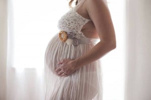 A pregnant person wearing a white lace and chiffon dress stands in front of a light source, holding their belly with one hand. The dress, adorned with a decorative floral belt, subtly hints at the beauty of embracing changes during pregnancy. The softly lit background adds an intimate touch.