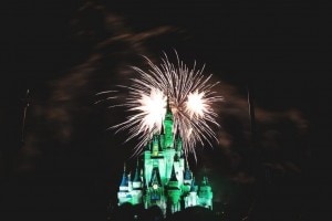 Fireworks burst over an illuminated castle at night, with green and white lights highlighting the castles towers against a dark sky.