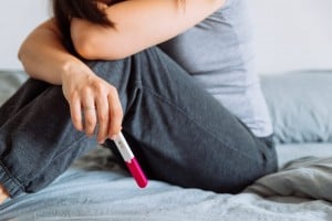 A person sits on a bed with their knees drawn up, resting their head on their arms, reflecting on their infertility battle. They are holding a pregnancy test with a pink cap in their hand. The person is wearing a grey shirt and dark grey pants.