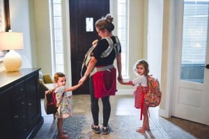 A woman standing by a front door holding a baby in a carrier, with multiple kids holding her hands. The children are wearing backpacks, and the scene is indoors with natural light coming through windows in the background. She's clearly getting out of the house with kids in tow.