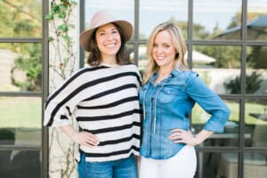 Two women stand smiling in front of a large window. One, possibly Esther Freedman, wears a striped blouse and a hat, while the other sports a denim shirt and white pants. They happily pose with one arm around each other in the sunny outdoor setting.