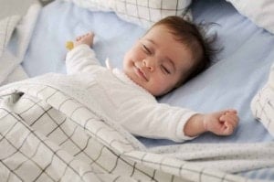 A baby lies on a bed, smiling with eyes closed and arms outstretched, covered by a white blanket with a black grid pattern.