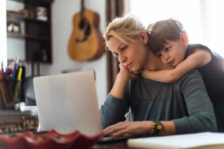 A woman is sitting at a desk working on a laptop with her hand resting on her chin, feeling like a bad mom. A young boy stands behind her with his arms wrapped around her shoulders, resting his head on her shoulder. A guitar hangs on the wall in the background.