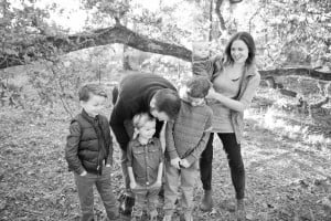 A family of six is outdoors in a wooded area. The parents stand with their four children; the mother holds the youngest, a baby, while smiling. The father leans down towards their three other young children, who are standing in front and looking down. They seem to be asking themselves if having another baby is on the horizon.