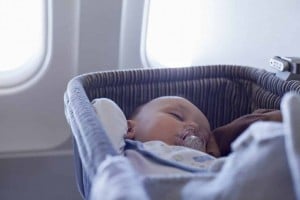 A baby sleeps in a bassinet with a pacifier in its mouth on an airplane, near two windows.