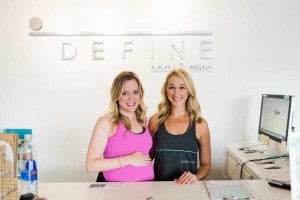 Two women stand at the reception desk of Define body & mind. The woman on the left, wearing a pink top, is pregnant and smiles warmly at the camera while resting her hand on her belly. The woman on the right, in a grey tank top, also beams at the camera.