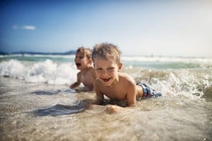 Two young boys lie in shallow ocean water at the beach, smiling and playing as small waves come in under a sunny sky.