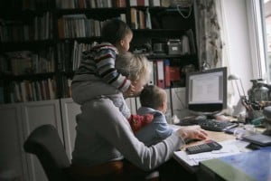 A mompreneur is working at a desk with a computer, holding a baby on her lap. Another child sits on her shoulders. The desk is cluttered with papers, a keyboard, and a calculator. Bookshelves filled with books are visible in the background.