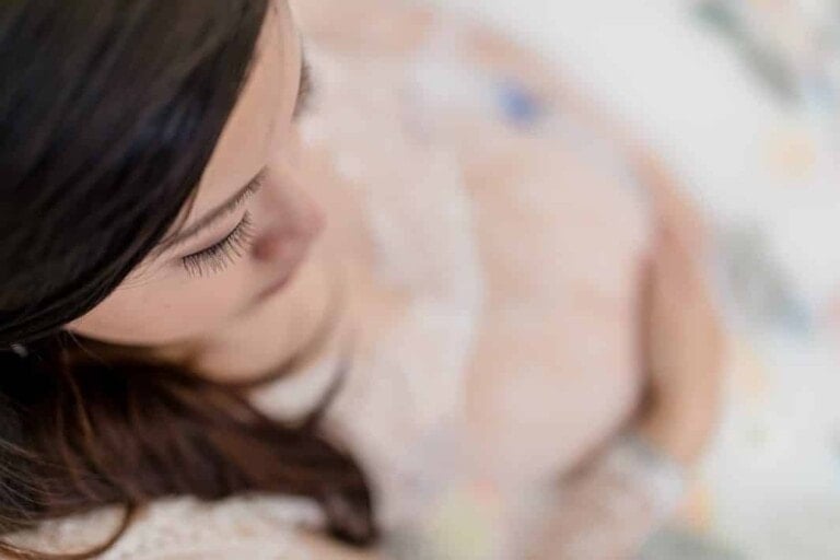 A close-up, top-down view of a person with long, dark hair, looking downward. The focus is on the person's eyelashes and head, while the background is blurred. The person appears to be wearing a light-colored, lace-like top, evoking the serene atmosphere of a maternity milk bath photoshoot.