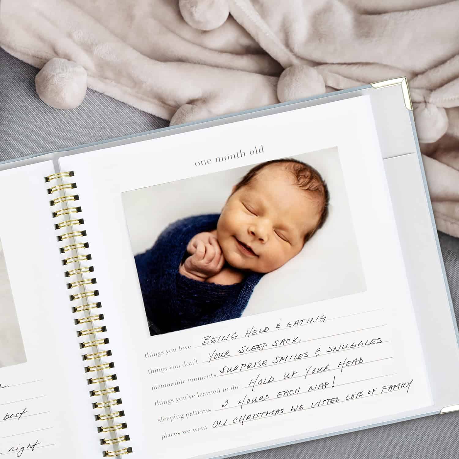 A scrapbook page features a photo of a newborn baby wrapped in a cozy blue blanket, smiling with eyes closed. The page, ideal as a DIY baby shower gift, is titled "one month old" with handwritten notes under headers like "favorite things to do" and "new things learned." Another handcrafted blanket peeks out.