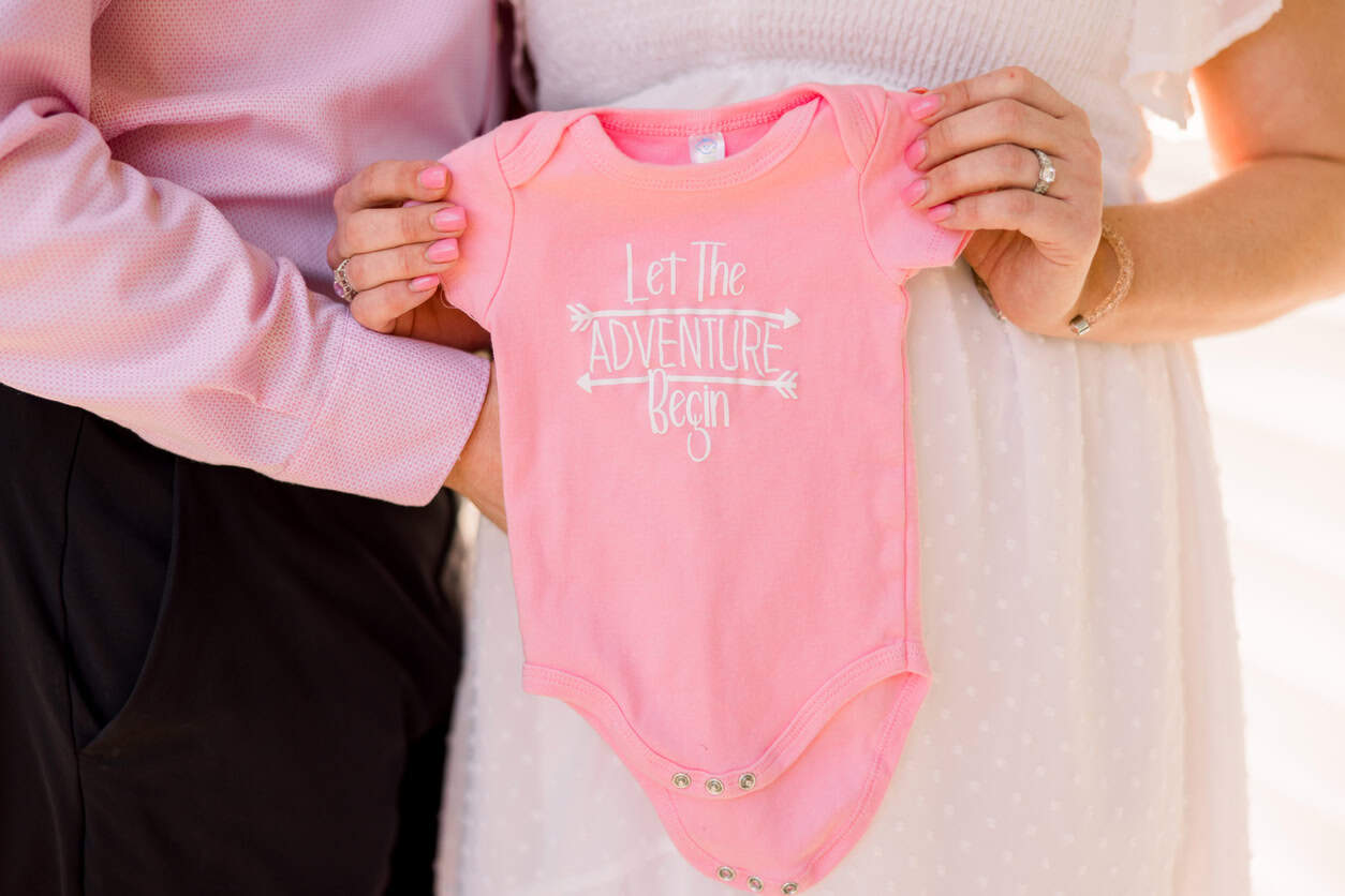 A couple holds up a pink baby onesie that reads, "Let The Adventure Begin." Their faces are out of the frame, and the woman's hand rests on her pregnant belly, emphasizing both the onesie and pregnancy. This heartfelt moment reflects their excitement for the journey ahead with a touch of diy baby gifts charm.
