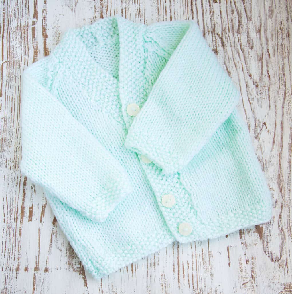 A light blue knitted baby cardigan with long sleeves and white buttons is displayed on a wooden surface, making it one of the perfect handmade baby gifts.