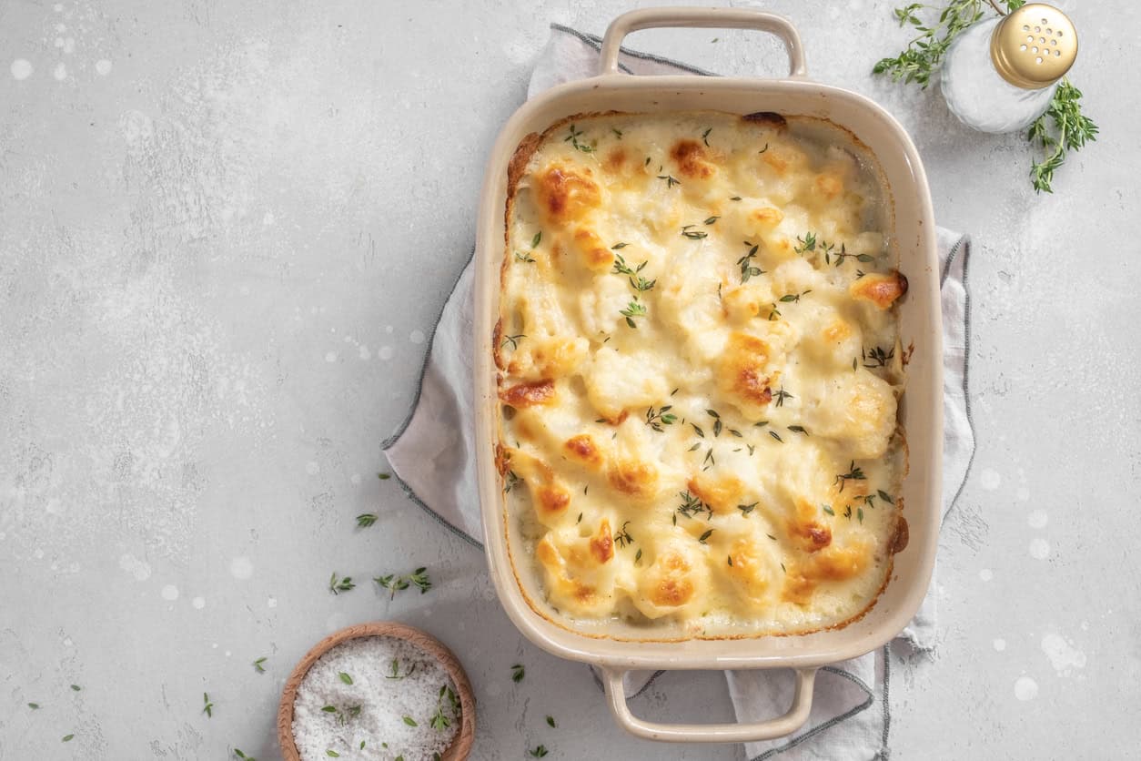 A rectangular baking dish filled with cooked cauliflower gratin topped with browned cheese and fresh thyme. Surrounding the dish are a gray cloth, a small bowl of salt, scattered thyme sprigs, and a salt shaker on a light gray surface. Perfect scenery for crafting diy baby shower gifts nearby!