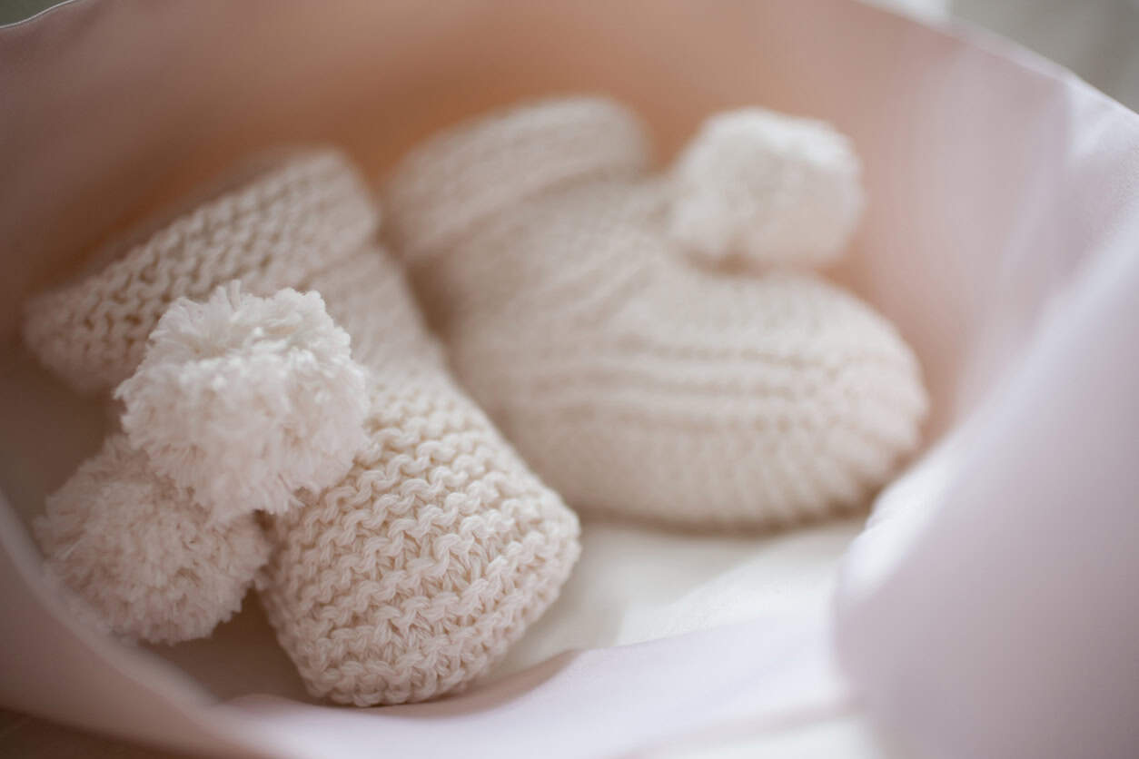 A close-up image of a pair of knitted baby booties in a soft, light beige color. Adorned with small pom-poms, these charming booties are the perfect homemade baby gift. They are placed on a light surface, partially wrapped in a light-colored cloth.