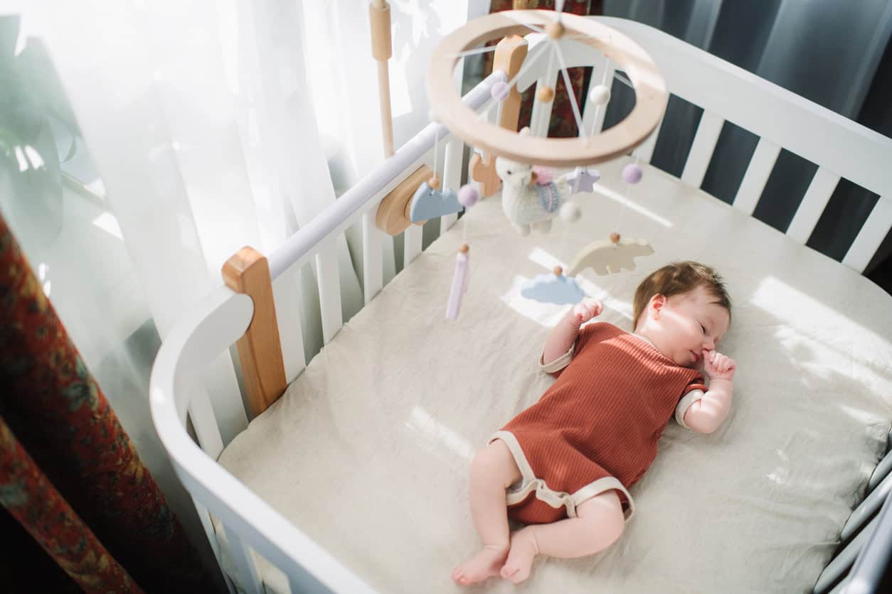 A baby in a brown onesie lies on their back in a white crib, with a mobile featuring various hanging items like clouds and stars positioned above. A heartfelt homemade baby gift, this DIY mobile captures the sunlight filtering through the curtain, illuminating the serene scene.
