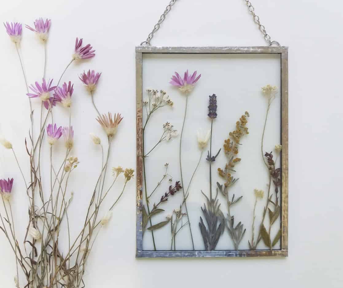 A decorative frame with pressed flowers hangs on a white wall, perfect for a homemade baby gift. To the left of the frame, a small bouquet of dried purple and white flowers is arranged vertically. The pressed flowers in the frame are displayed in a variety of colors and species.