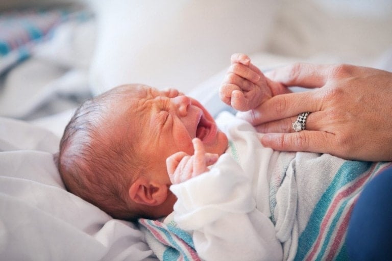 A newborn baby is crying while lying on a white bed. An adult's hand, wearing a ring, is gently holding the baby's hand. The baby, wrapped in a striped blanket and wearing a white outfit, might have conditions like tongue tie that can make feeding challenging.