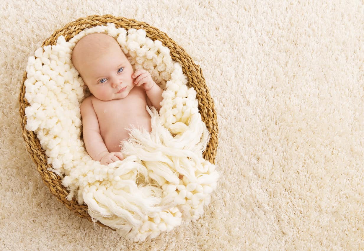 A baby is lying in a woven basket lined with a soft, cream-colored blanket. The baby is looking off to the side, wrapped snugly in the blanket, and resting on a beige, textured surface—the perfect scene for memorable handmade baby gifts.