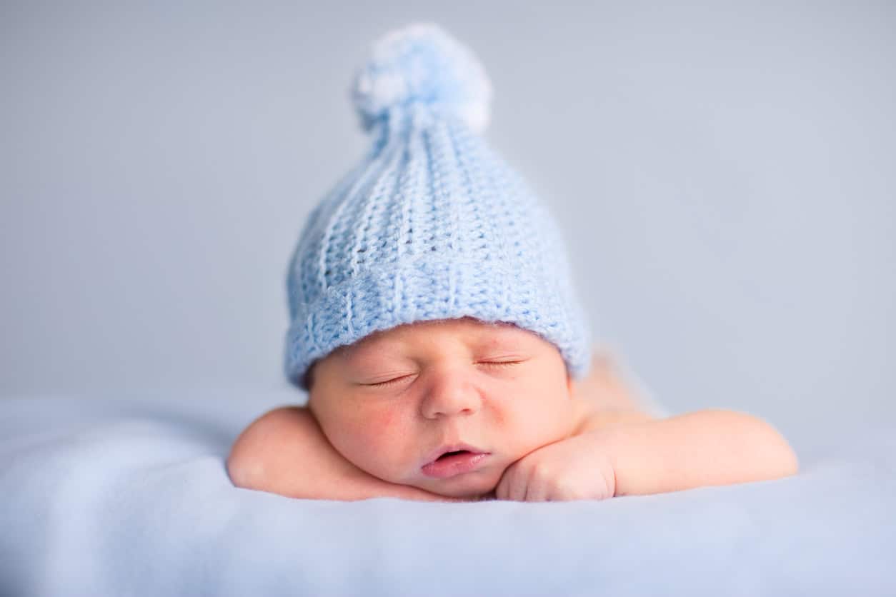 A newborn baby wearing a light blue knitted hat with a pom-pom is sleeping on its stomach with its head resting on its arms. The background, also light blue, creates a soft and peaceful atmosphere—perfect for inspiring handmade baby gifts or unique DIY baby shower gifts.