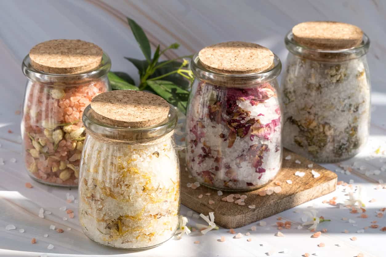Image of four glass jars with cork lids containing different types of bath salts. The jars are placed on a wooden board, each with a distinct mixture of pink salts, dried flowers, and herbs. Green plant leaves are in the background, making these perfect handmade baby gifts for any special occasion.