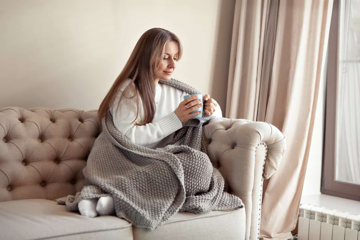 A woman with long hair sits on a beige tufted sofa, wrapped in a chunky gray blanket. Holding a mug with both hands, she gazes down at it thoughtfully. Light filters in through a large window with beige curtains in the background, perhaps pondering her next DIY baby shower gift idea.