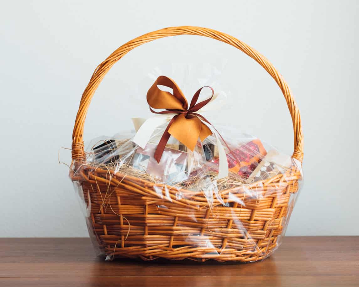 A wicker gift basket wrapped in clear plastic and adorned with a brown and white ribbon sits on a wooden surface. Perfect for DIY baby shower gifts, the basket contains various items, partially obscured by the wrapping.