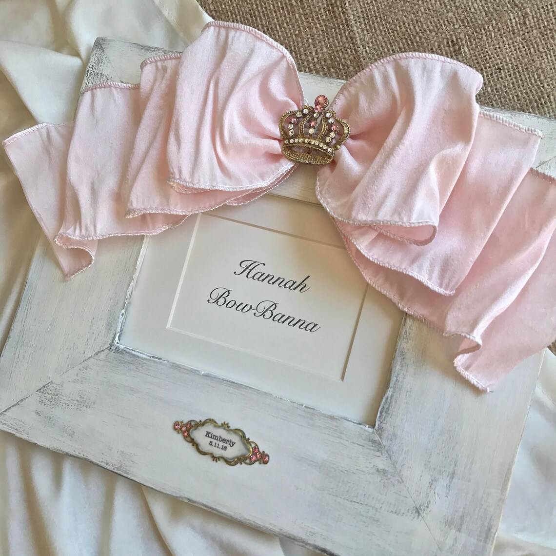 A white picture frame adorned with a large, pink, satin bow and a small decorative crown in the center of the bow. The frame contains a placeholder card that reads "Hannah Bow-Bowanna". Perfect as a DIY baby shower gift, the frame lies on a beige fabric background.