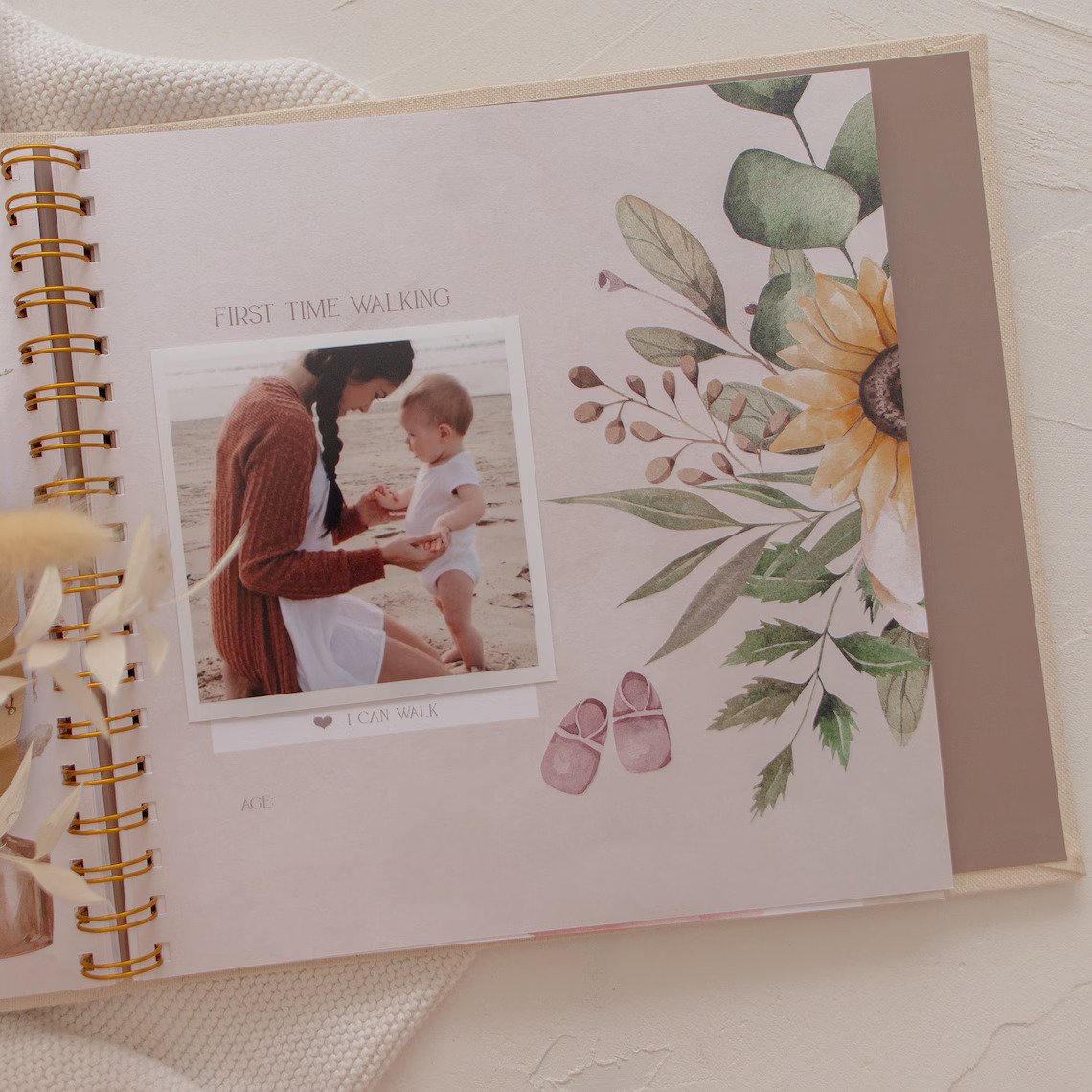 A scrapbook page featuring a photograph of an adult and a baby labeled "First time walking." The page, highlighting the joy of DIY baby gifts, has floral designs, a sunflower, and illustrations of baby shoes. The adult is guiding the baby who is standing on a sandy surface.