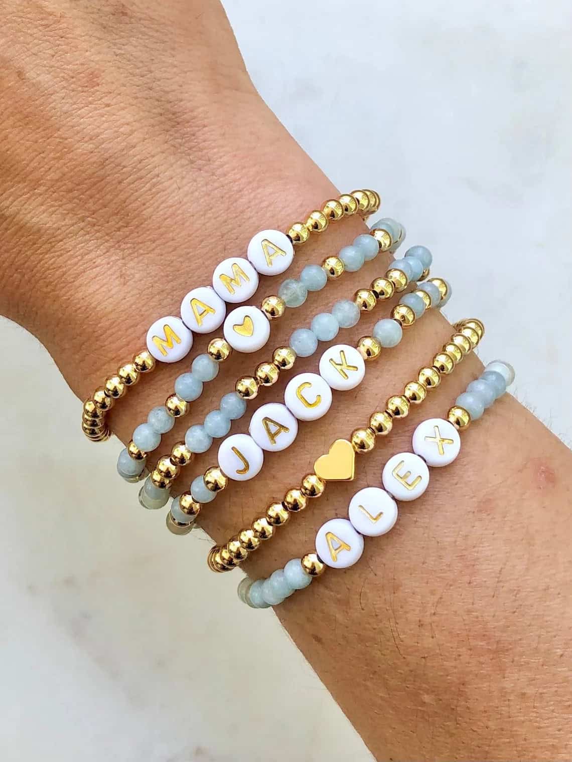 A wrist is adorned with four beaded bracelets, perfect as a DIY baby shower gift. Each bracelet has white beads spelling out "MAMA," "JACK," and "ALEX," interspersed with gold and light blue beads. A small heart-shaped bead separates the names, adding a sweet touch to this personalized keepsake.