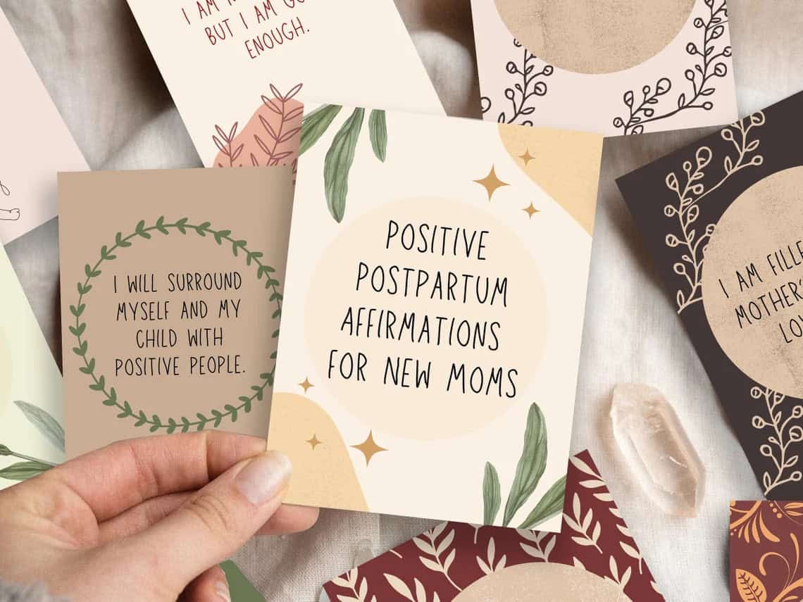A person holds a card with the text "I will surround myself and my child with positive people." Other cards with various positive affirmations for new moms are spread around, including one that reads "Positive Postpartum Affirmations for New Moms." Natural leaves, crystals, and lovely handmade baby gifts complete the serene scene.