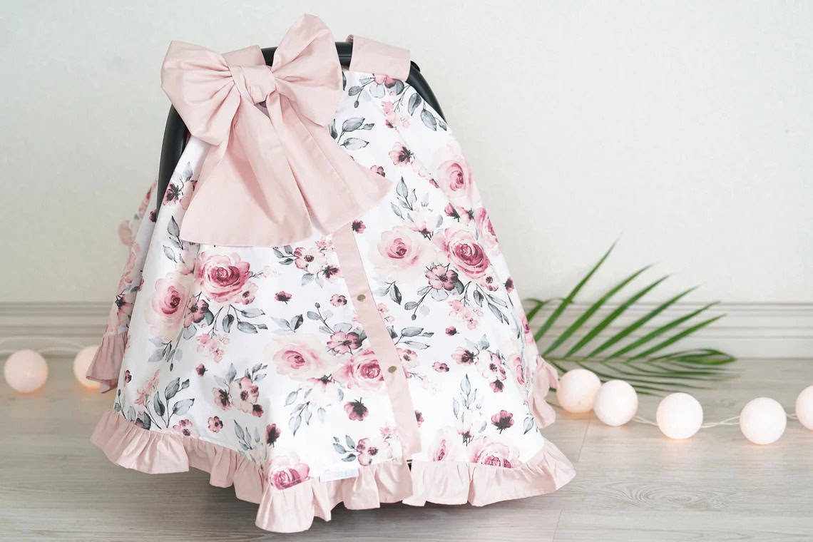 A floral baby car seat cover with pink and white roses and a large pink bow is draped over a car seat. The cover has a light pink ruffled hem. Nearby are a palm leaf and string lights with round white bulbs. This adorable setup makes for perfect DIY baby shower gifts, set against a gray background.