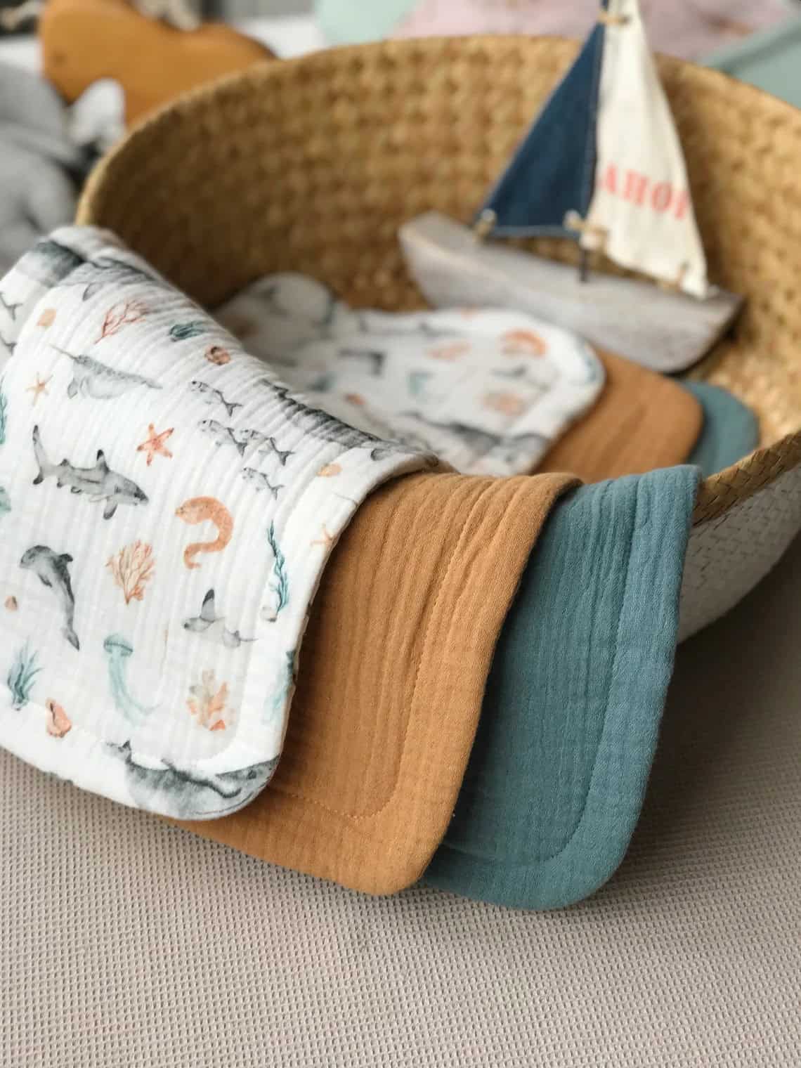 A wicker basket contains three swaddle blankets folded over its edge. The blankets are in white with sea-themed prints, mustard yellow, and teal colors. In the background, a small decorative sailboat is also inside the basket, making it a charming choice for homemade baby gifts.