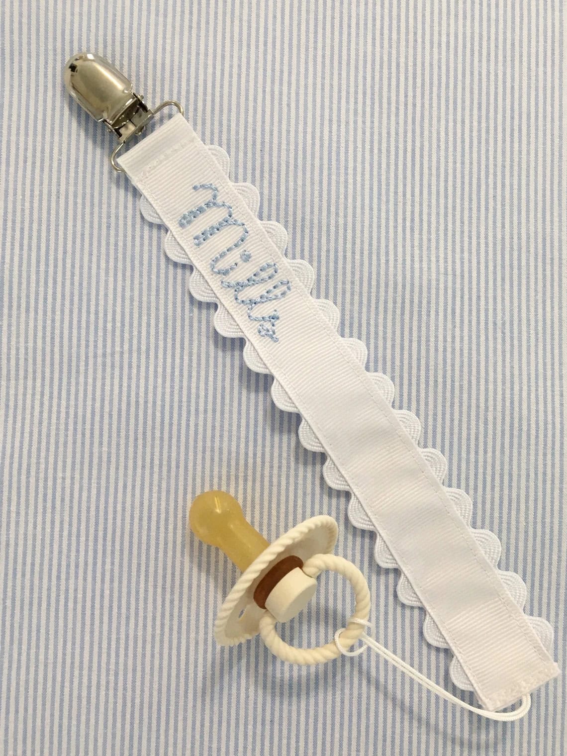 A pacifier with a beige rubber nipple attached to a white clip-on strap, perfect for DIY baby gifts. The strap is embroidered with a name in blue thread and has a scalloped edge, set against a charming blue and white striped fabric background.